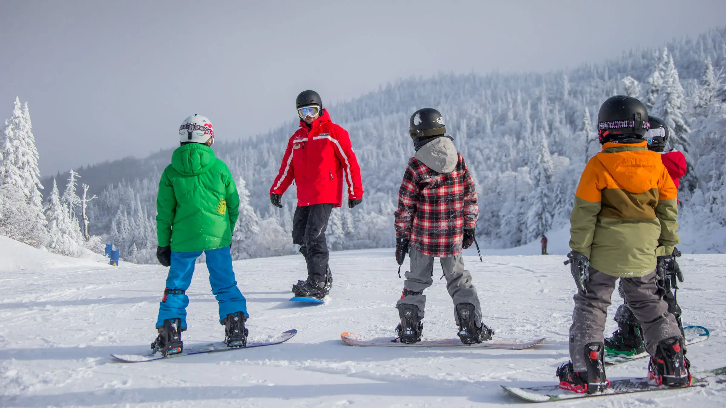 Skupina dětí na snowboardech s instruktorem v červené bundě, připravující se na trénink na zasněženém svahu.