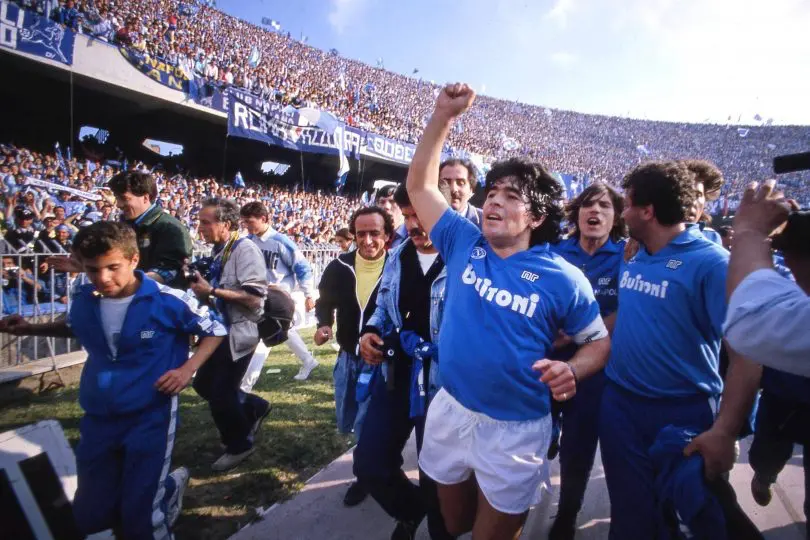 Maradona se zvednutou rukou slaví triumf s modrým dresem a fanoušky Neapole, kteří ho zbožňují.