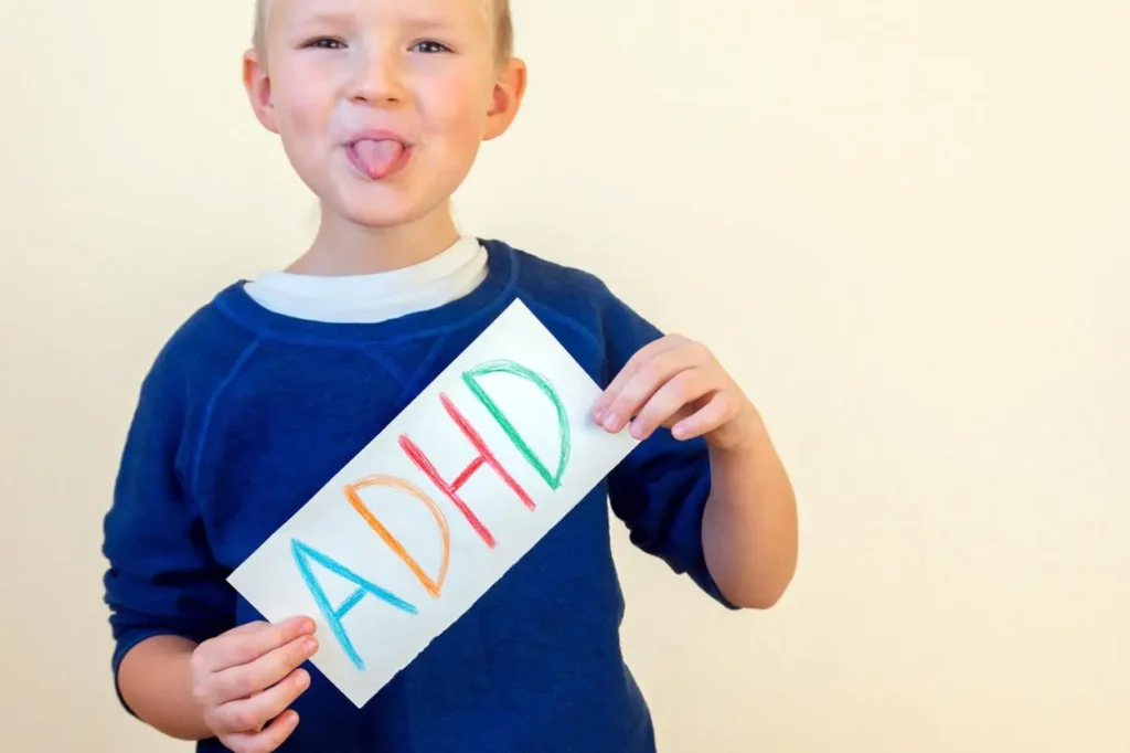 Chlapec s krátkými blond vlasy v modré mikině drží v ruce cedulku s nápisem „ADHD“, která je napsána barevnými písmeny. Vyplazuje jazyk a vypadá hravě a rozverně.