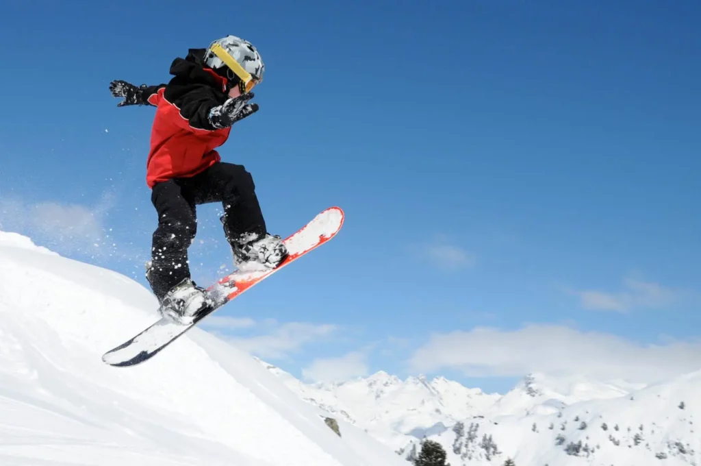 Dítě na snowboardu ve skoku na zasněženém kopci, s modrou oblohou a horskou krajinou v pozadí.