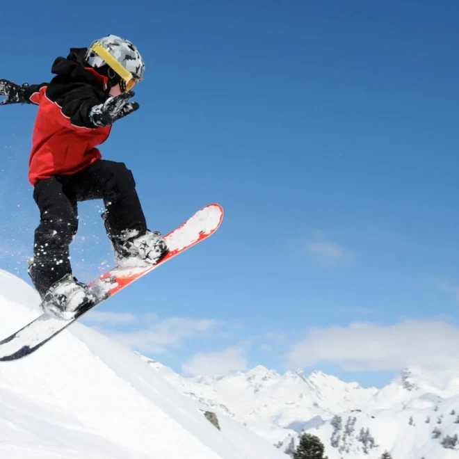 Dítě na snowboardu ve skoku na zasněženém kopci, s modrou oblohou a horskou krajinou v pozadí.