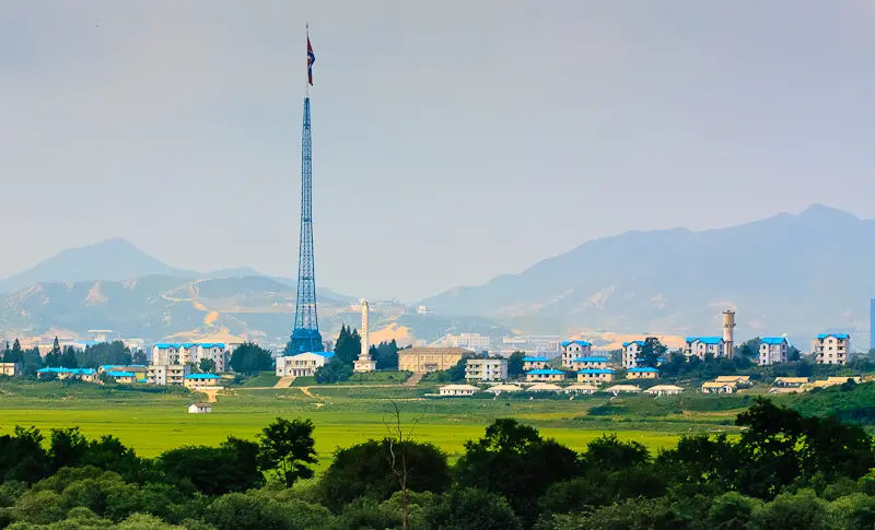 Pohled na Kijŏng-dong, ukazující jasné barvy budov a rozsáhlou krajinu kolem, s dominantní vlajkou Severní Koreje na vysokém stožáru. Hory v pozadí dotvářejí atmosféru.