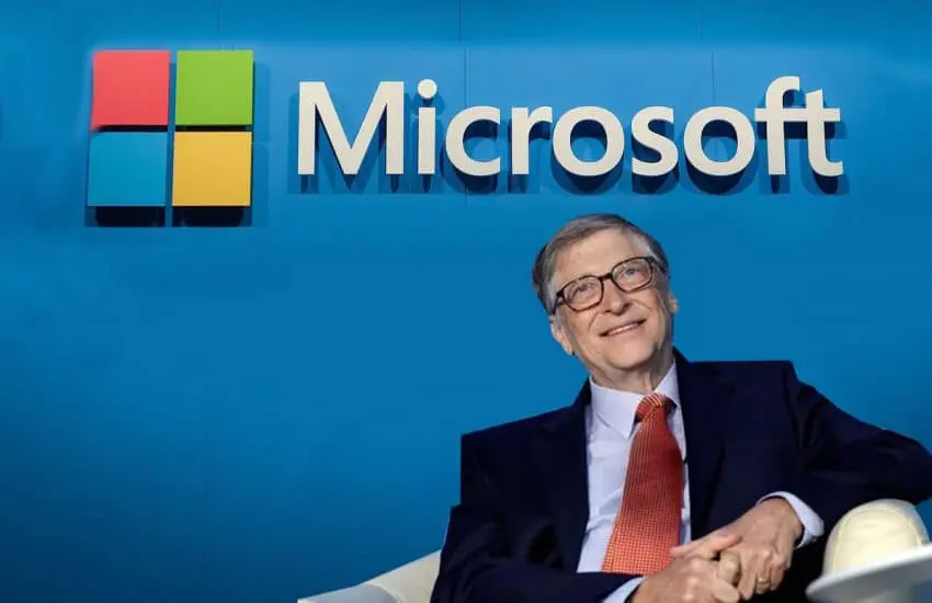 Bill Gates sedící před logem společnosti Microsoft, usmívající se. Zakladatel Microsoftu v obleku a kravatě na modrém pozadí.