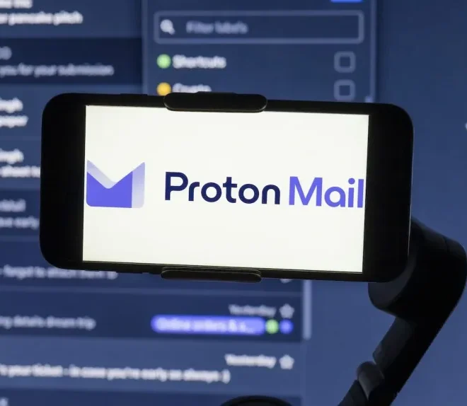 Smartphone zobrazující logo Proton Mail, s rozostřeným náhledem e-mailového rozhraní v pozadí.