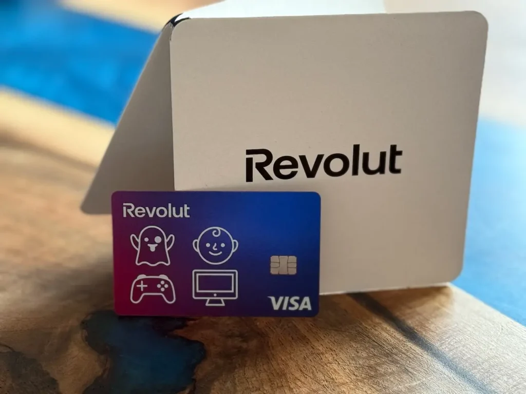 Revolut platební karta s hravými ikonami položená před elegantním bílým obalem s logem Revolut, zachyceno na stylovém dřevěném stole.