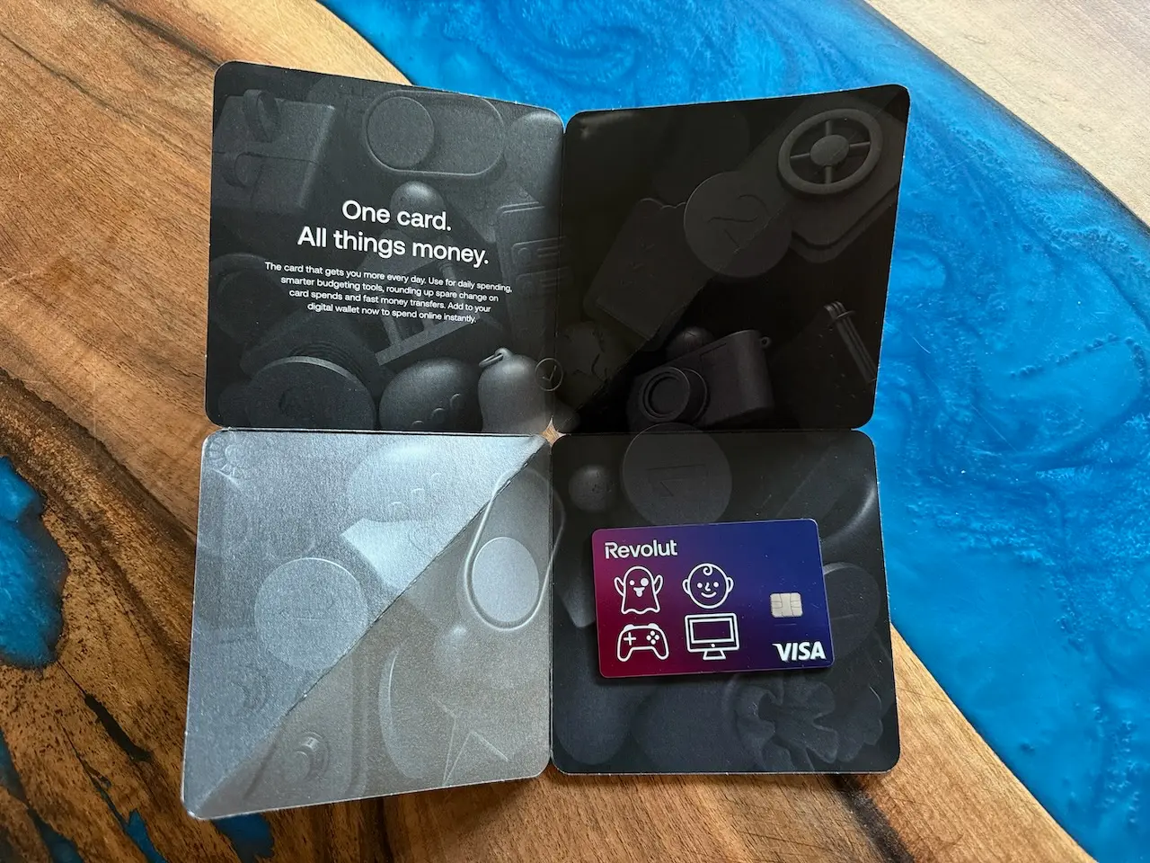 Revolut platební karta v otevřeném obalu s moderním designem a nápisem „One card. All things money.“ na dřevěném stole s modrou pryskyřicí.