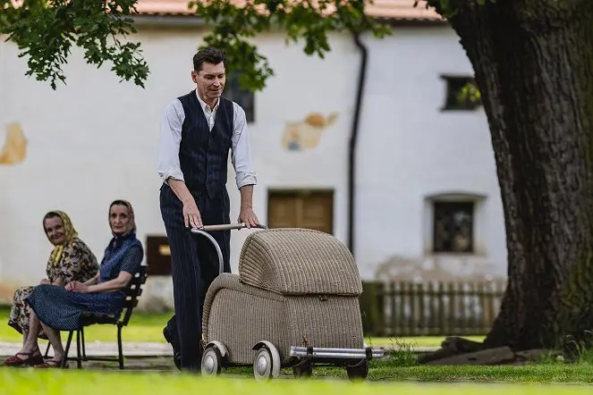  David Švehlík v elegantní vestě tlačí proutěný retro kočárek na travnatém prostranství, zatímco na lavičce sedí dvě starší ženy a sledují jej s úsměvem.