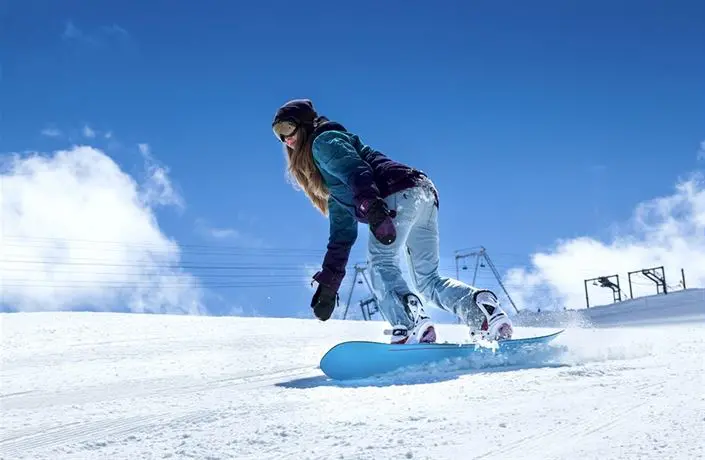 Snowboardistka sjíždí zasněžený svah na modrém snowboardu za slunečného počasí, v pozadí lanovky a modrá obloha.