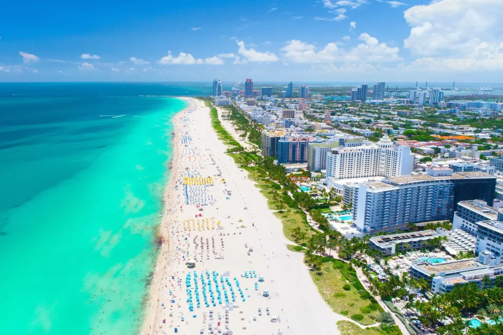 Letecký pohled na slavnou pláž South Beach v Miami na Floridě s tyrkysovým oceánem, bílým pískem a řadami barevných slunečníků, obklopenou vysokými hotely a městskými budovami.