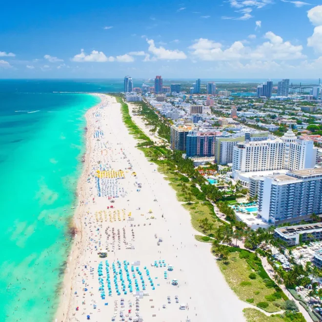 Letecký pohled na slavnou pláž South Beach v Miami na Floridě s tyrkysovým oceánem, bílým pískem a řadami barevných slunečníků, obklopenou vysokými hotely a městskými budovami.