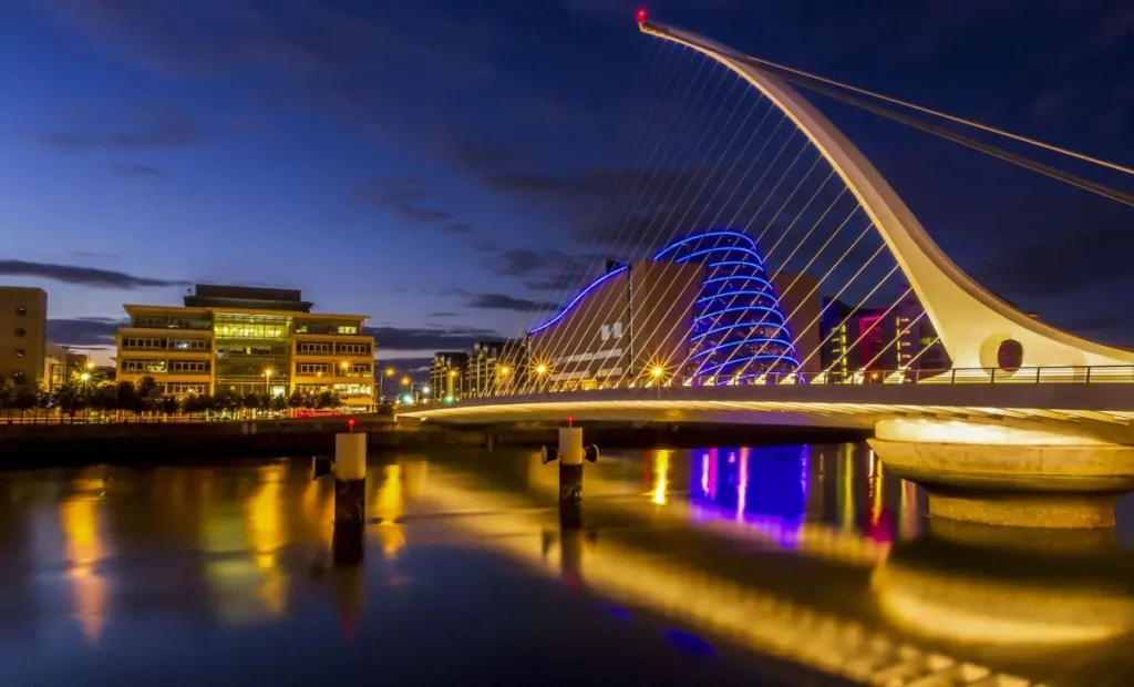 Noční pohled na ikonický most Samuel Beckett Bridge v Dublinu, Irsko, s nádherně osvětleným Kongresovým centrem Dublin v pozadí, jehož modré a fialové světlo se odráží na hladině řeky Liffey.