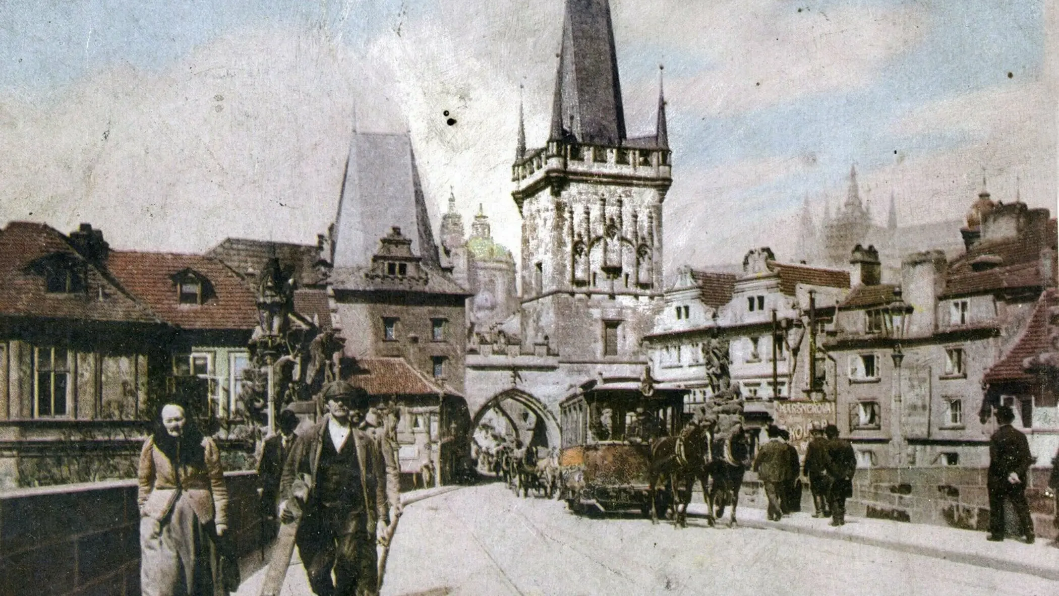 Historická fotografie Karlova mostu s pohledem na Staroměstskou mosteckou věž a Pražský hrad v pozadí, zachycující život na mostě v minulých stoletích.