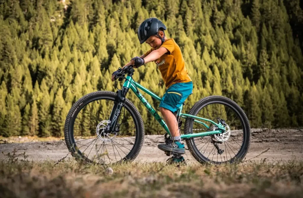 Dítě na horském kole projíždí terénem obklopené lesem. Kolo je vybaveno odpruženou vidlicí a hydraulickými kotoučovými brzdami, ideální pro mladé cyklisty.