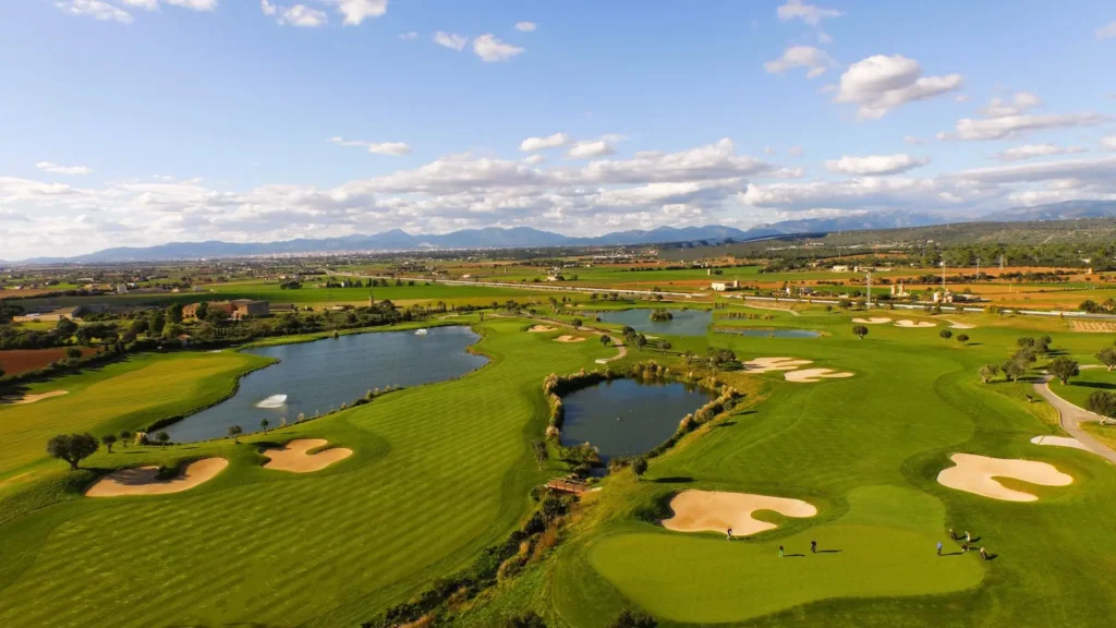 Letecký pohled na jedno z nejlepších golfových hřišť na Mallorce s rozlehlými greeny, vodními překážkami a okolní přírodou v pozadí s horským panoramatem.