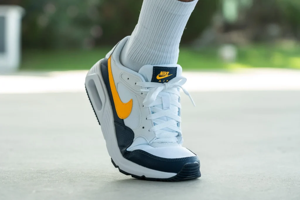 Bílé a tmavě modré tenisky Nike Air Max s oranžovým logem Swoosh na noze člověka s bílými ponožkami, focené venku na betonovém povrchu.