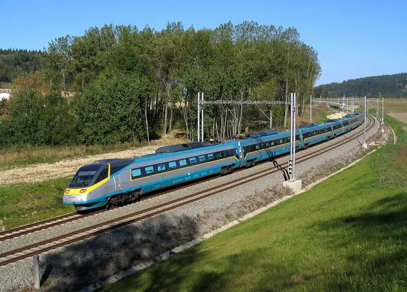 Pendolino vlak v přírodě na trati obklopené stromy a zelení, projíždějící otevřenou krajinou.