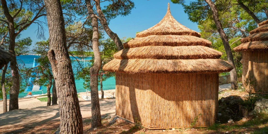 Rákosový bungalov v Pine Beach Resortu Pakoštane u moře obklopený borovicemi, s výhledem na azurové moře a plachetnici.