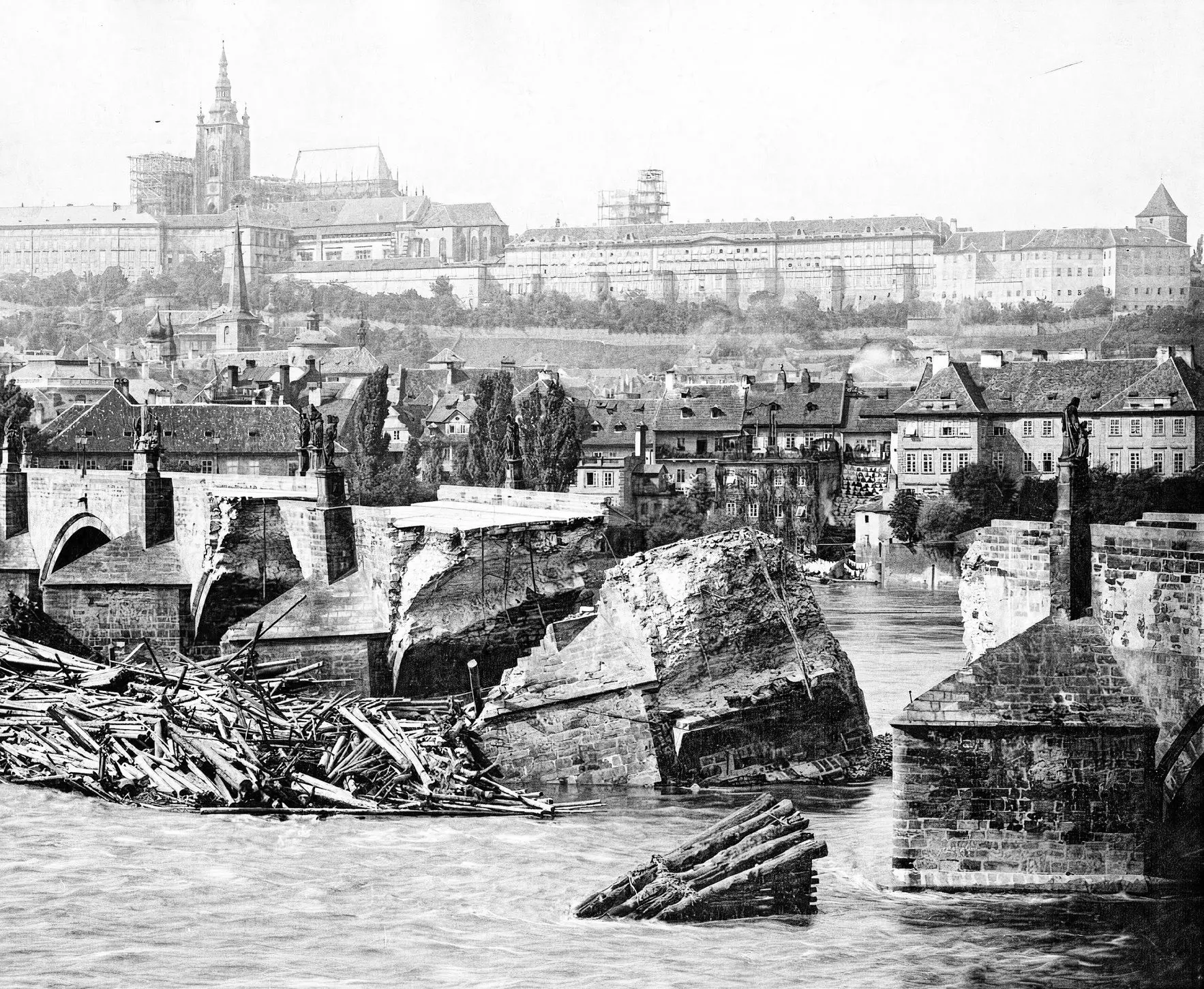 Černobílá fotografie zničených pilířů Karlova mostu během povodně v roce 1890, s troskami dřeva zachycenými u pilířů.