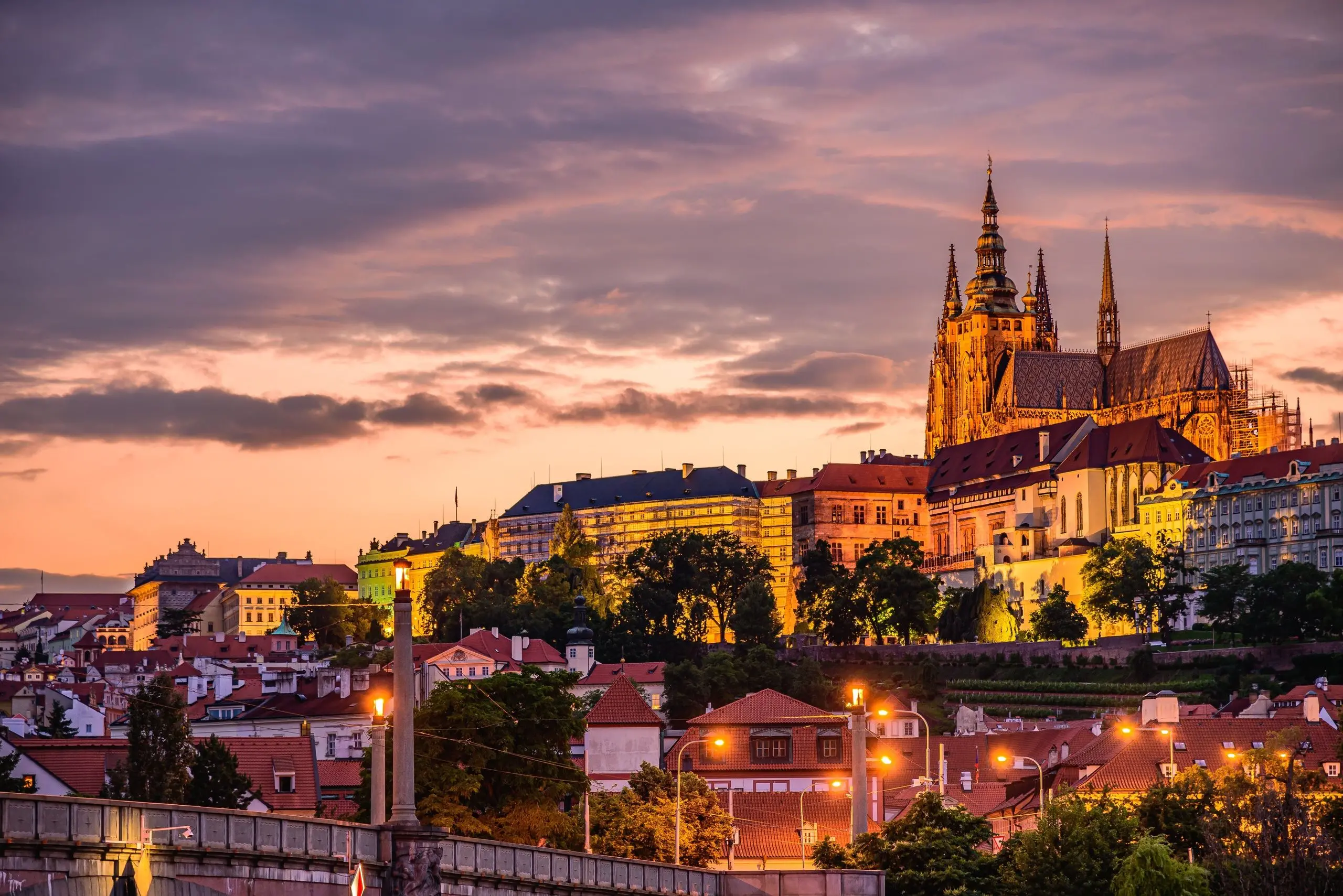 Noční pohled na Pražský hrad a osvětlenou katedrálu sv. Víta s dramatickým západem slunce nad Prahou.