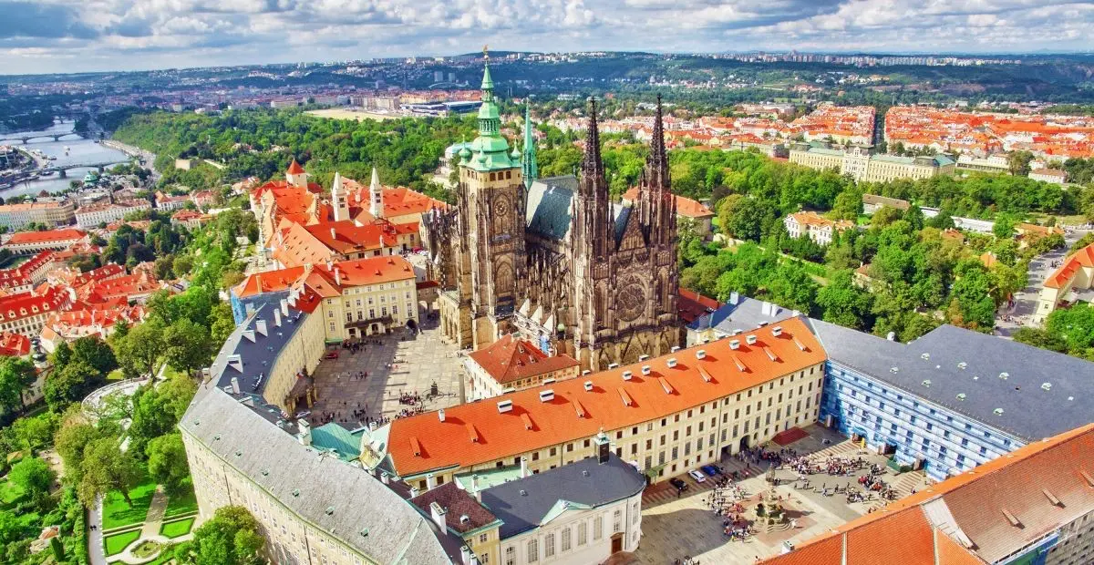 Letecký pohled na Pražský hrad s katedrálou sv. Víta, nádvořími a zahradami obklopenými historickými budovami a panoramatem města.