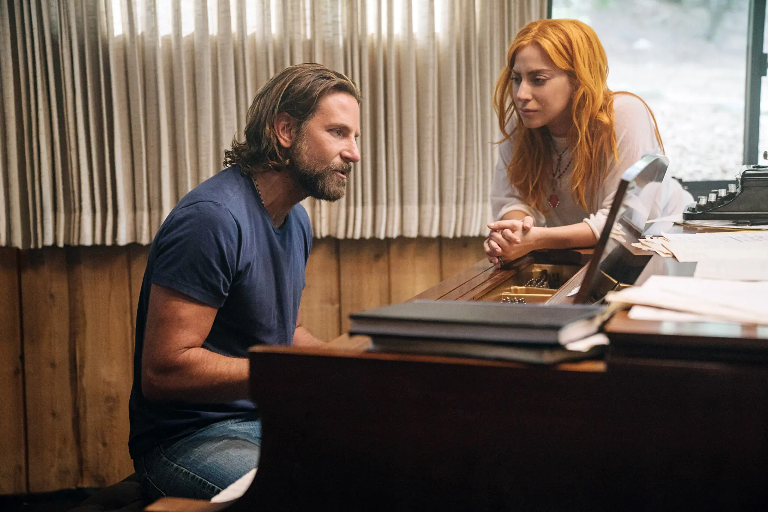 Bradley Cooper (Jackson Maine) hraje na klavír, zatímco Lady Gaga (Ally) ho pozorně poslouchá a opírá se o klavír. Intimní a inspirativní scéna ze studia plná hudební kreativity.