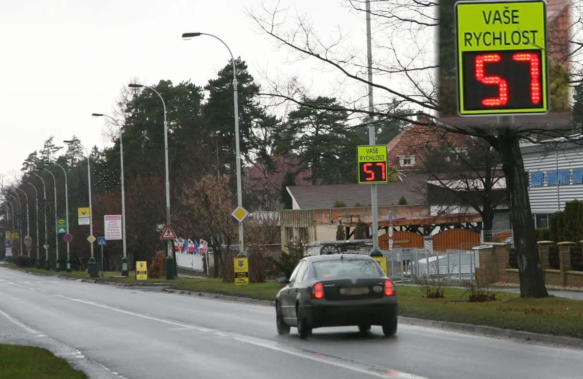 Automobil projíždějící kolem měřicího radaru s displejem zobrazujícím rychlost 57 km/h na silnici v obci.