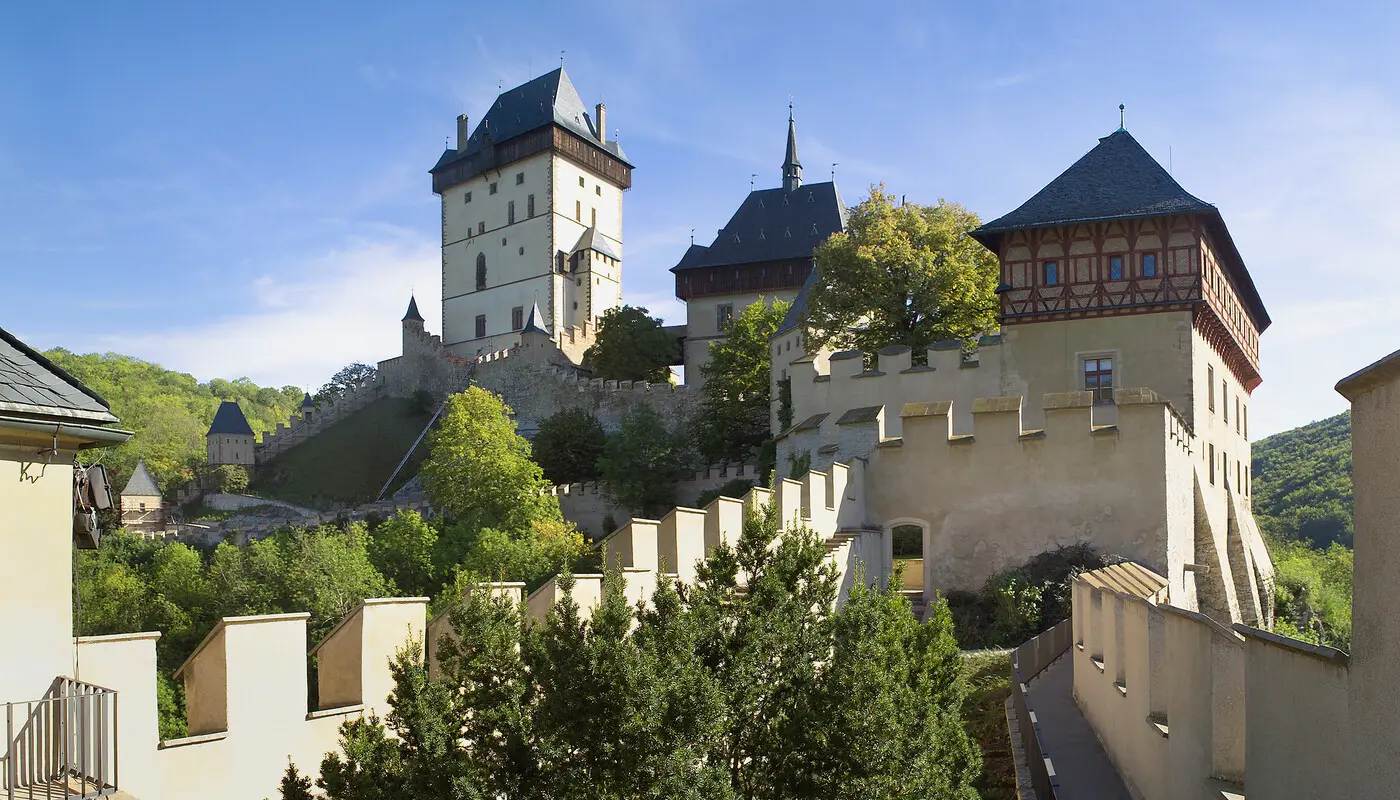 Pohled na hrad Karlštejn s opevněním a centrální Velkou věží, zasazený do zeleného údolí.