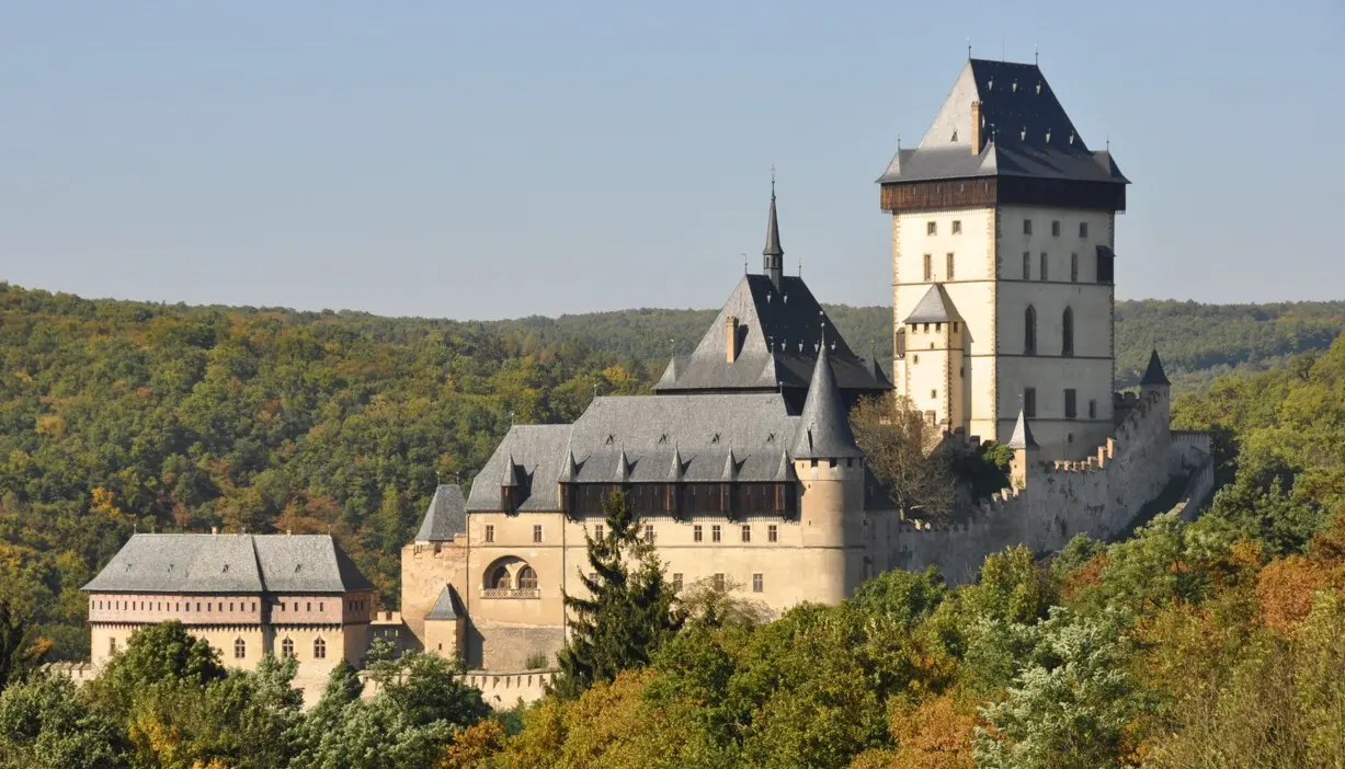 Celkový pohled na Karlštejn obklopený lesy, zachycující hradby, věže i gotické střechy.