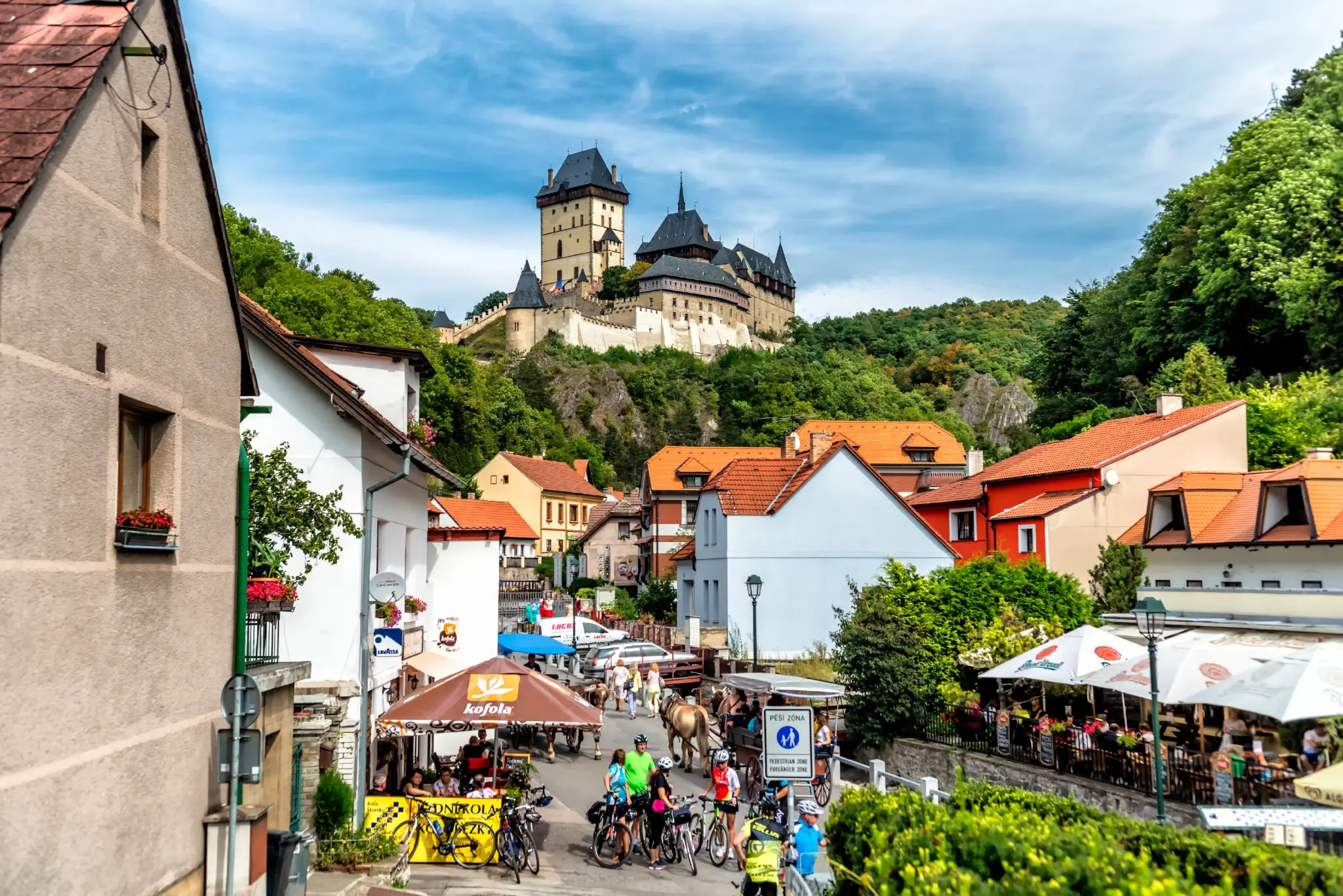 Rušná ulička vedoucí k hradu Karlštejn s turisty, kavárnami a výhledem na hrad v pozadí.