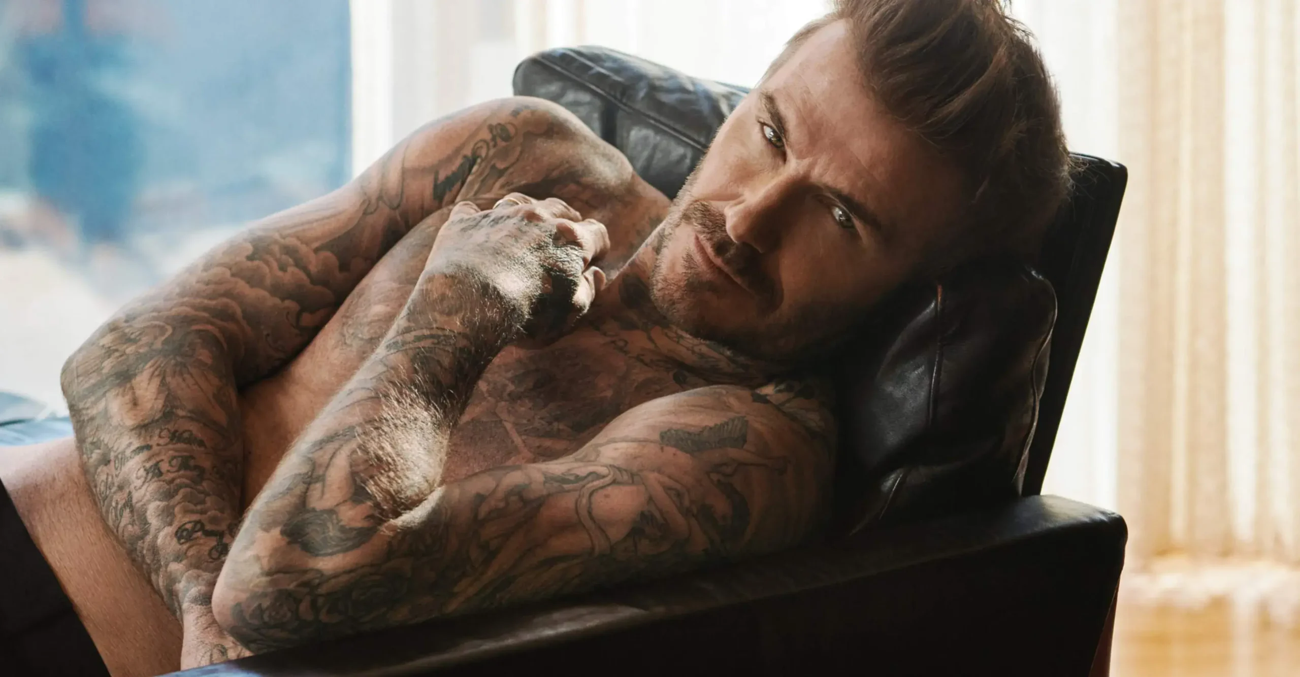 Charismatický David Beckham leží na černém koženém křesle, odhalující své tetování a hledící přímo do kamery.