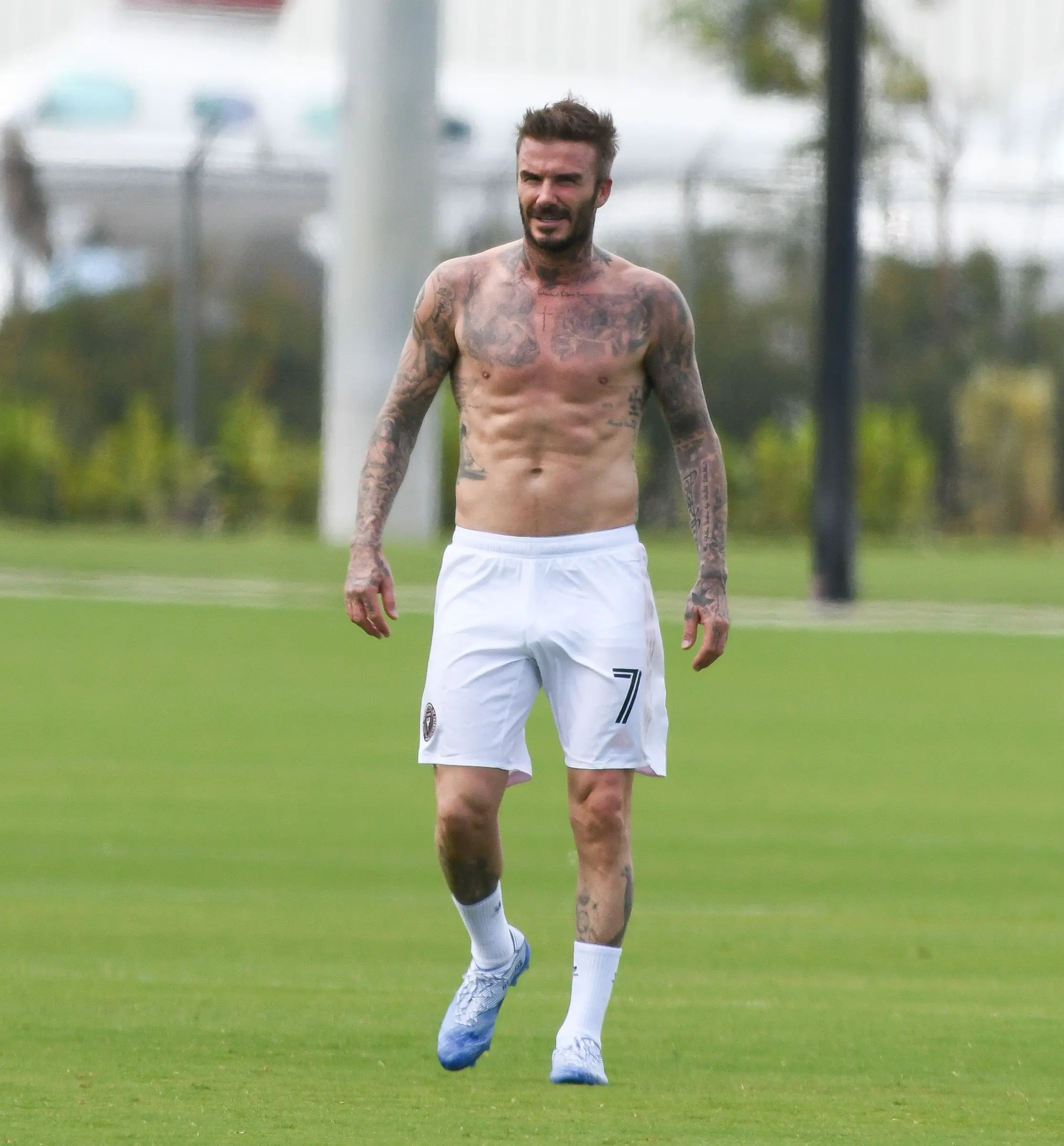 Bývalý fotbalista David Beckham běží po zeleném trávníku v bílých šortkách s číslem 7, odhalující svou atletickou postavu a rozsáhlé tetování.