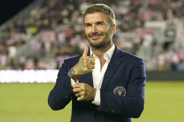 David Beckham v elegantním saku s logem Inter Miami CF na stadionu, ukazující palec nahoru s úsměvem na tváři. V pozadí je vidět publikum s růžovými prvky.