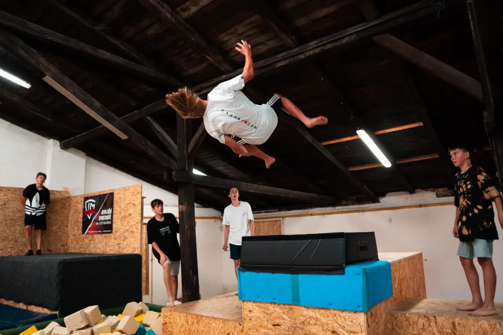 Mladý sportovec provádí akrobatický backflip v kryté parkourové hale, zatímco ostatní sledují jeho skok.