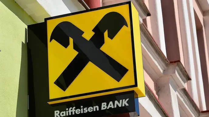 Logo Raiffeisenbank na žluté ceduli s černými zkříženými kladivy, umístěné na fasádě budovy.