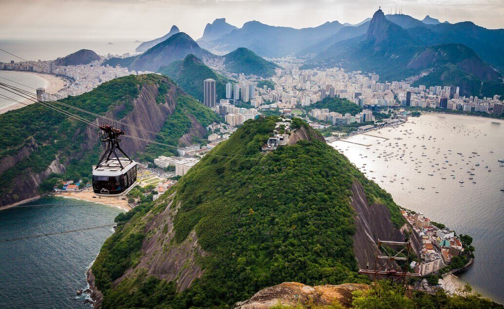 Panoramatický pohled na Rio de Janeiro s ikonickou lanovkou na Cukrovou homoli, zelenými kopci a městem rozprostřeným podél pláží a zátoky.