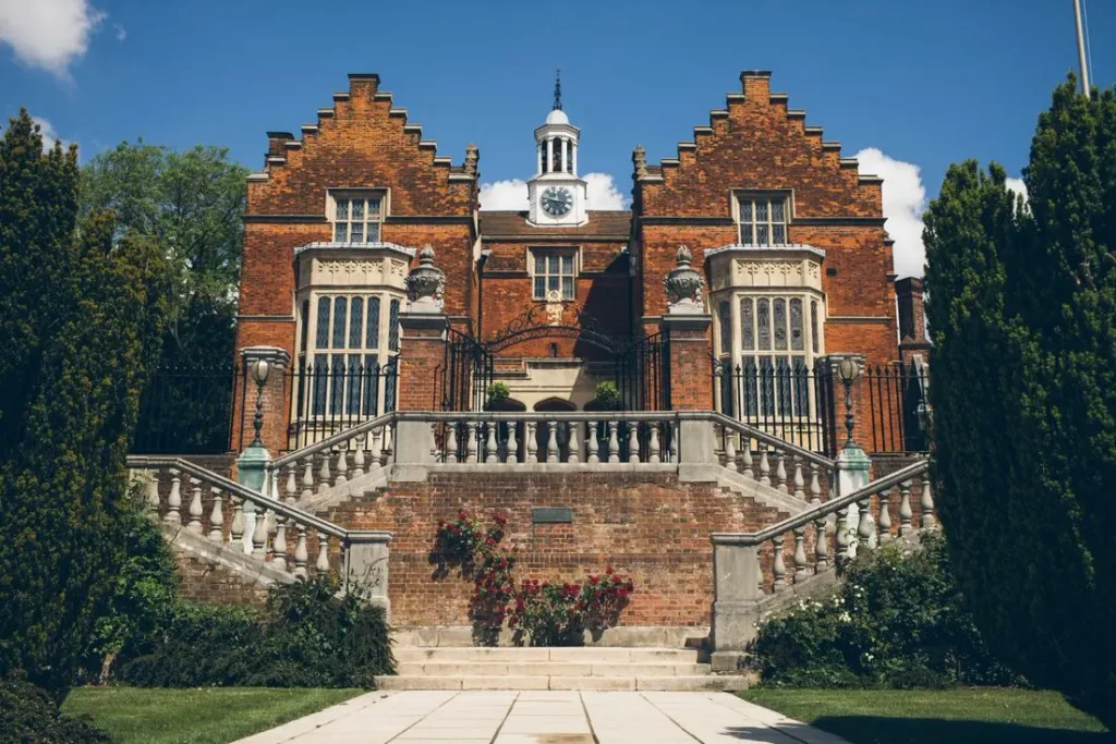 Pohled na historickou budovu Harrow School v Anglii s červenou cihlovou fasádou, ozdobnými schody a zelení v okolí.