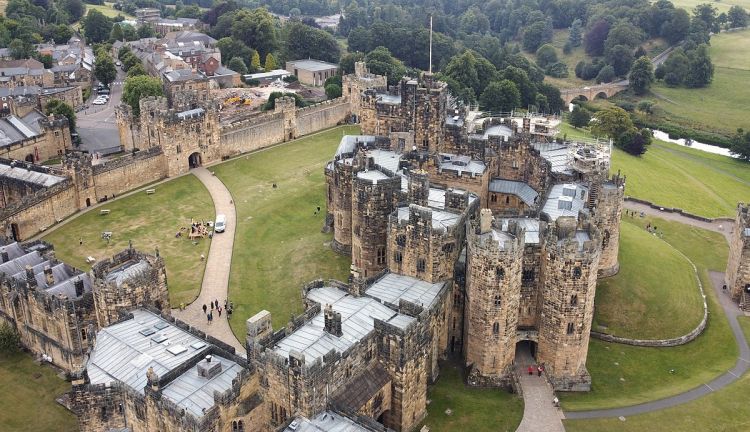 Letecký pohled na hrad Alnwick v Anglii s mohutnými kamennými zdmi, věžemi a okolní zelenou krajinou.