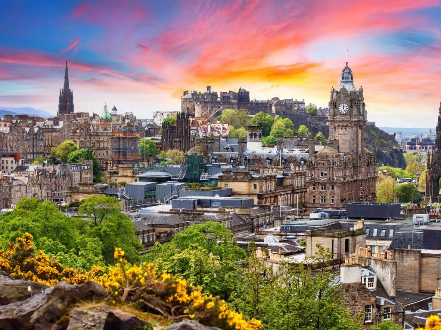 Panoramatický pohled na historické centrum Edinburghu při západu slunce, s dominantou Edinburského hradu na kopci a krásně zbarvenou oblohou.