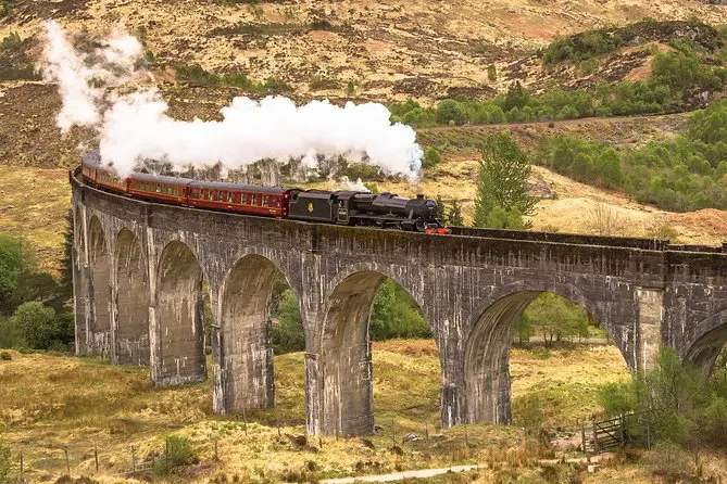 Slavný parní vlak Jacobite přejezd přes ikonický Glenfinnan Viaduct ve Skotsku, obklopený zelenou krajinou, známý také z filmů o Harrym Potterovi.