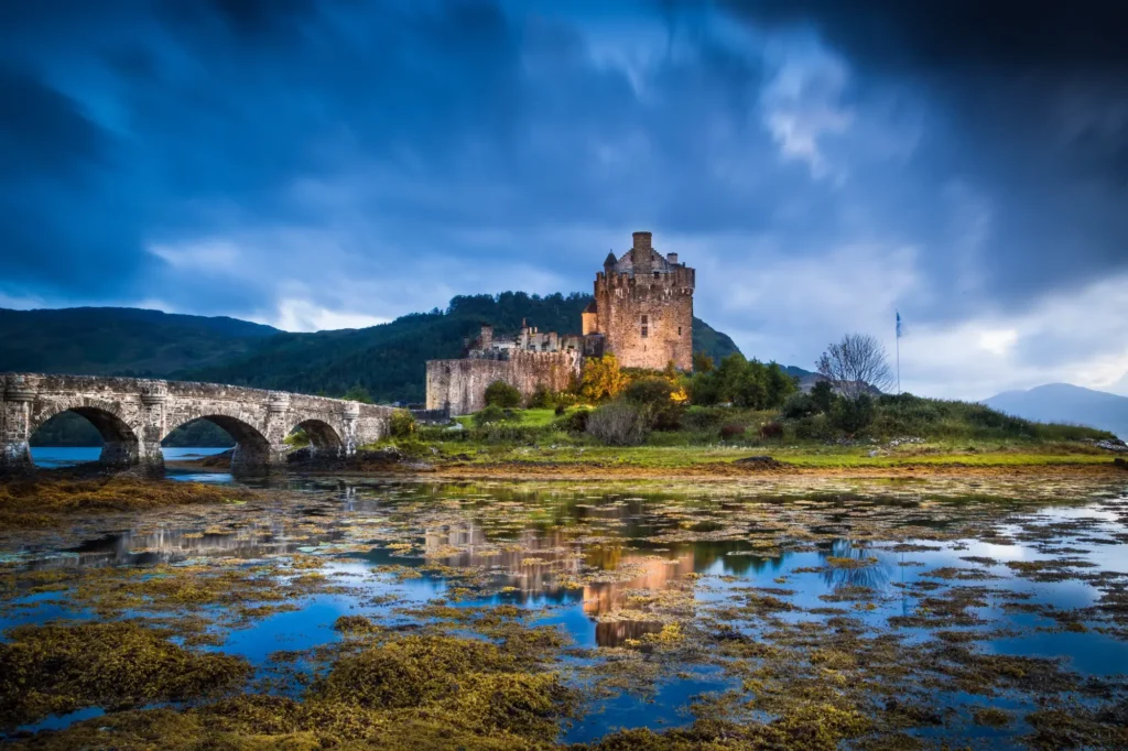 Historický skotský hrad Eilean Donan, majestátně stojící na ostrově obklopeném vodou, s kamenným mostem vedoucím k pevnosti a dramatickou oblohou v pozadí.